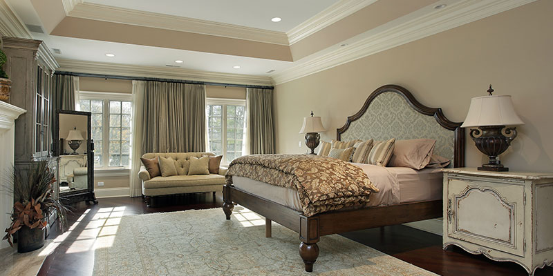 Bright bedroom with tray ceiling, recessed lights, and seating by a sunny window.