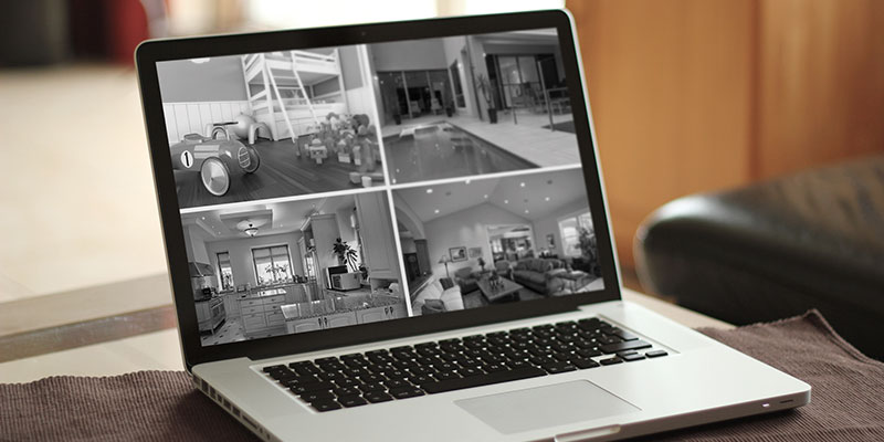 Open laptop displaying a four-camera home security feed (playroom, pool area, entryway, and living room) on a table in a living room.