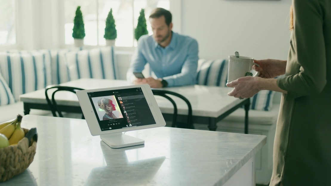 Tabletop touchpad on a kitchen island shows music playing through the Control4 multiroom audio system as a woman walks past it to go to the dining table. 