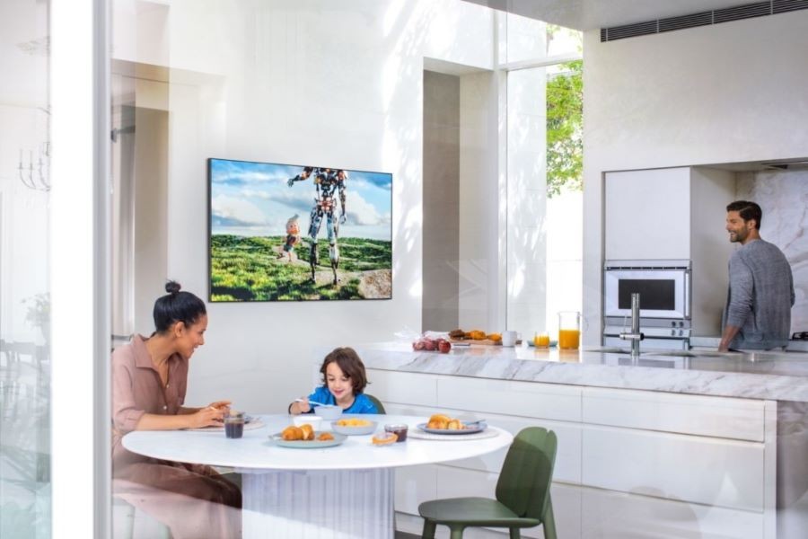 A family eating breakfast in the kitchen with a TV display on the wall.