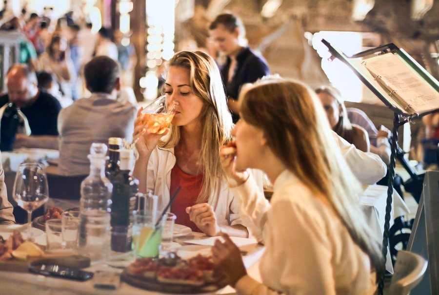 Diners sitting at a table in a busy restaurant, a woman drinking out of a glass of white wine.